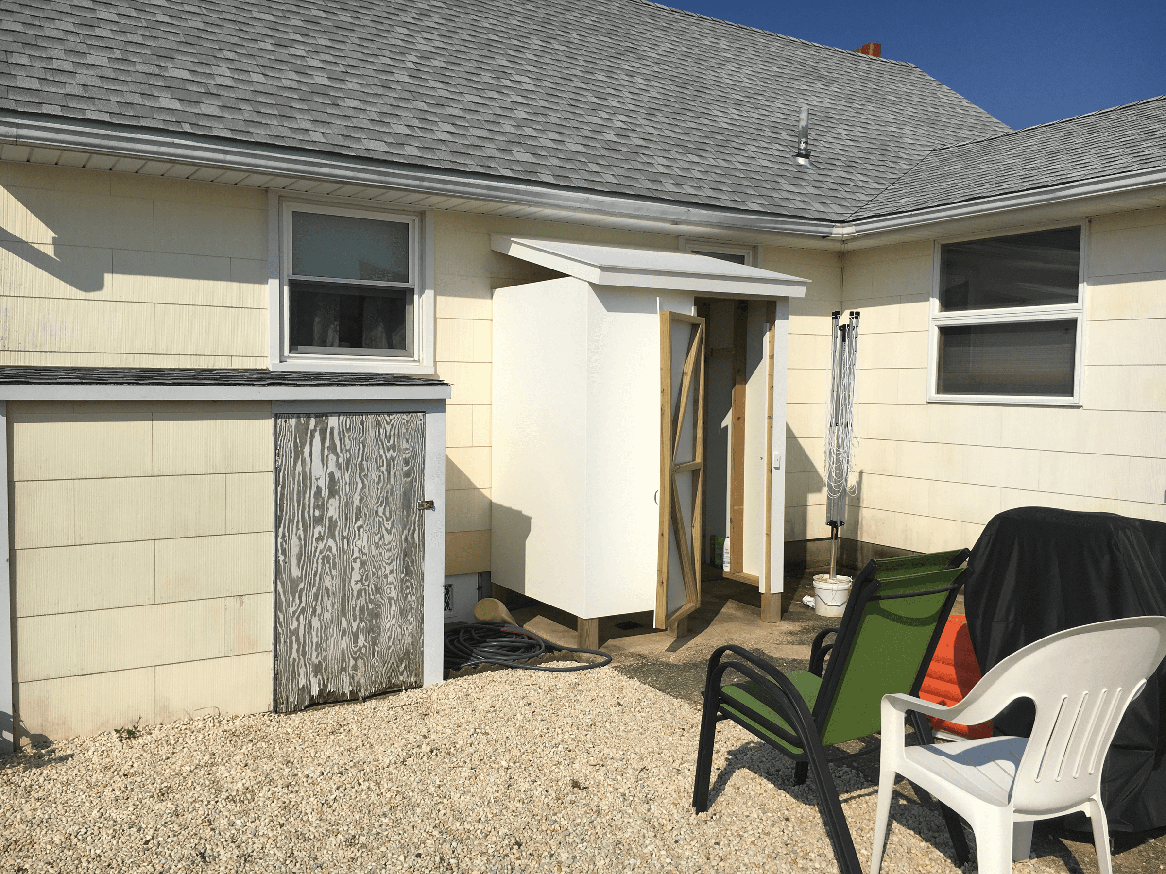 Back corner of a house with cream siding and a white shed, before painting