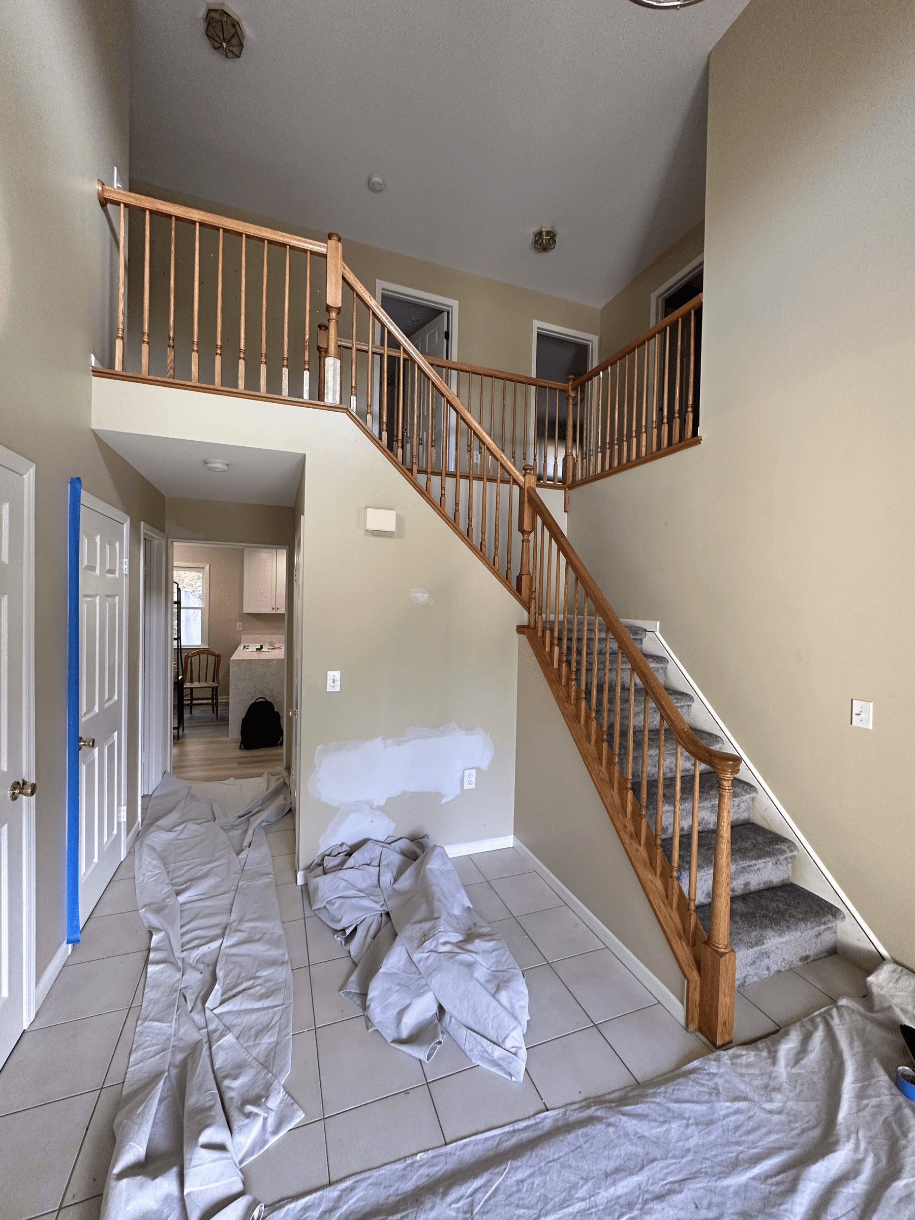 Two-story foyer and staircase with beige walls and the floor covered in drop cloths, before painting